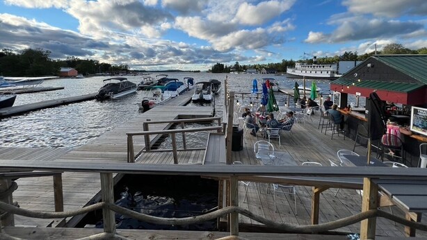 A wooden pier with boats docked on the water, a bar area with outdoor seating, and a small building with a green roof. - Lakeside Loft (Direct Access to Moosehead Lake!) (Greenville)