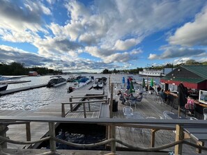 A wooden pier with boats docked on the water, a bar area with outdoor seating, and a small building with a green roof.