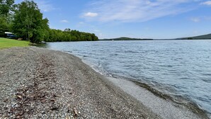 A pebbled beach with a calm lake, surrounded by dense green trees and a clear sky. - Lakeside Loft (Direct Access to Moosehead Lake!) (Greenville)