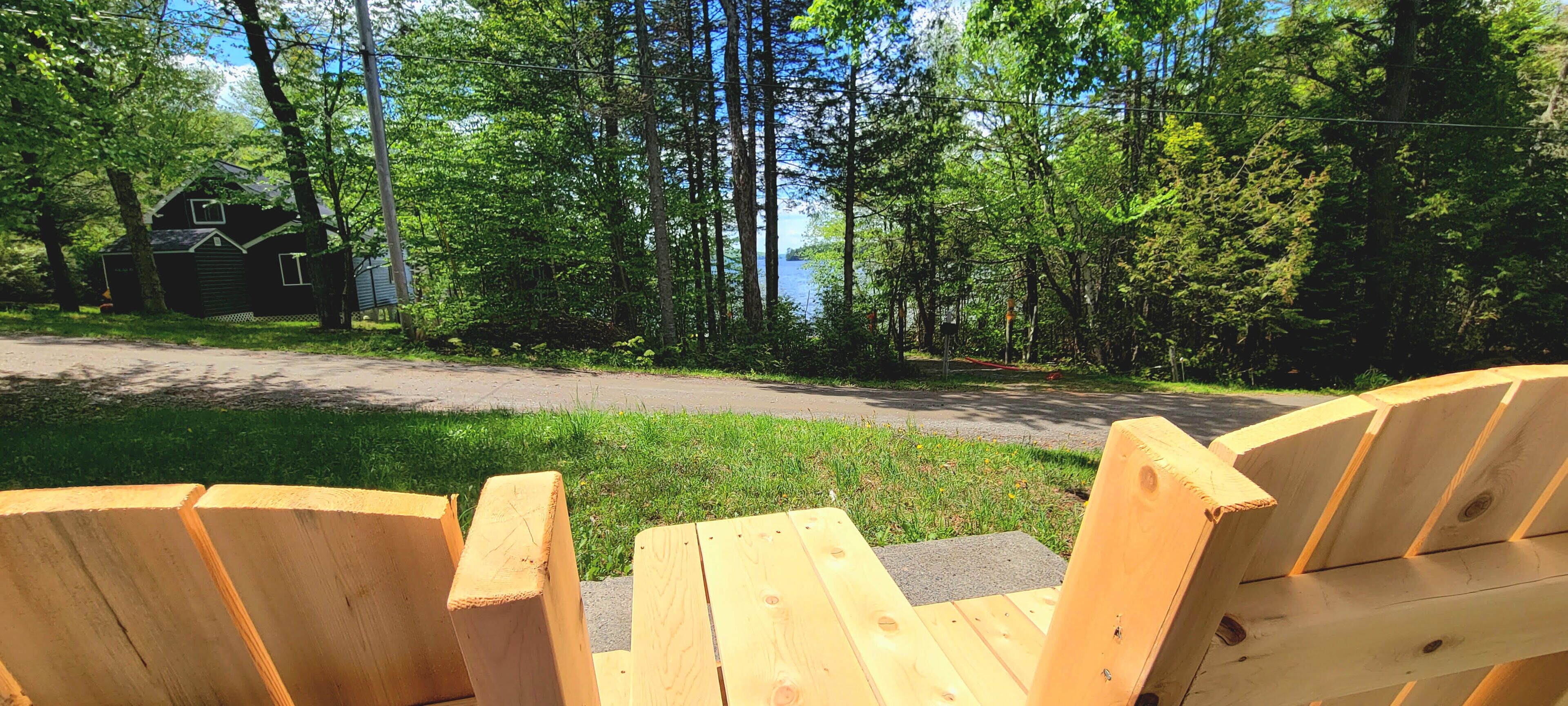A wooden deck with Adirondack chairs overlooking a road with a house and dense forest.