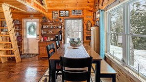 Ein Blockhaus-Interieur mit einem Holzespeisetisch, Stühlen, einer Himmelsleiter, einem Bücherregal und einem großen Fenster mit Blick auf Schnee.