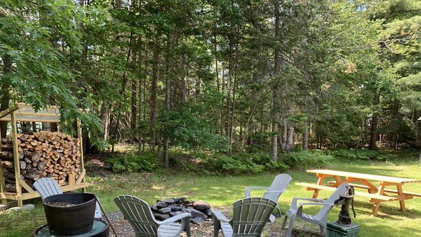 A rustic outdoor seating area with Adirondack chairs, a fire pit, and a picnic table surrounded by a forest. - Tranquil Times (In Town with Direct ATV/Snowmobile Access!) (Greenville)