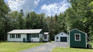 A rustic cabin with a gravel driveway leading to a green shed and a white building with a metal roof. - Tranquil Times (In Town with Direct ATV/Snowmobile Access!) (Greenville)