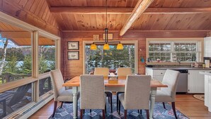 L’intérieur d’un chalet en bois comprend une table à manger, des chaises, un coin cuisine et de grandes fenêtres donnant sur des arbres et un lac.