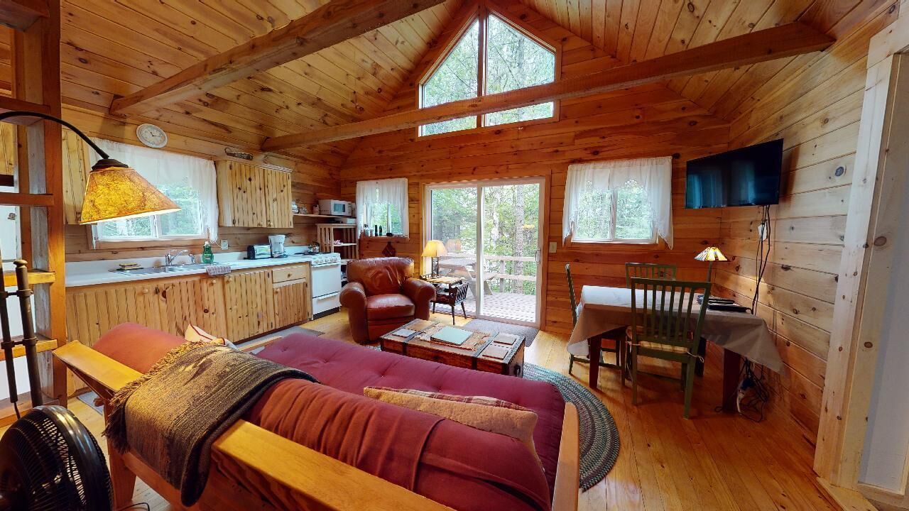 A cabin interior with a wooden ceiling, a sofa, a dining table, a TV, and a kitchen area.