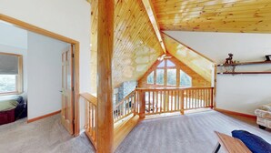 Una stanza con soffitto spiovente, travi in legno, camino in pietra, un balcone con ringhiera in legno e vista sugli alberi.
