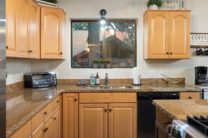 A kitchen with wooden cabinets, a built-in dishwasher, a stove, and a window with a view of trees and a brick building. - Cozy Munds Park Cabin ~ Fire Pit & Sunset Views (Munds Park)
