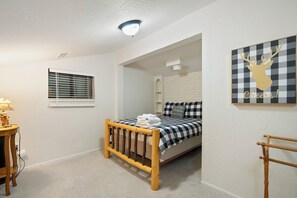 Ein Schlafzimmer mit einem Bett, einem Fenster mit Jalousien, einer Lampe, einem Wandspiegel und einem gerahmten Bild.