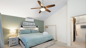 A bedroom with a bed, bedside table, lamp, wall-mounted air conditioner, and a ceiling fan. - Cozy Munds Park Cabin ~ Fire Pit & Sunset Views (Munds Park)