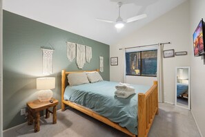 A bedroom with a bed, a nightstand, a wall-mounted macrame decoration, a ceiling fan, a window with a view, and a framed inspirational quote. - Cozy Munds Park Cabin ~ Fire Pit & Sunset Views (Munds Park)