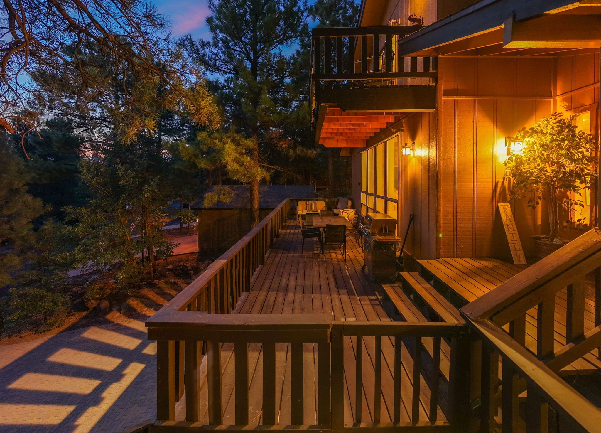 Une terrasse en bois avec des sièges extérieurs, entourée d’arbres et bordée d’un bâtiment doté d’un balcon.