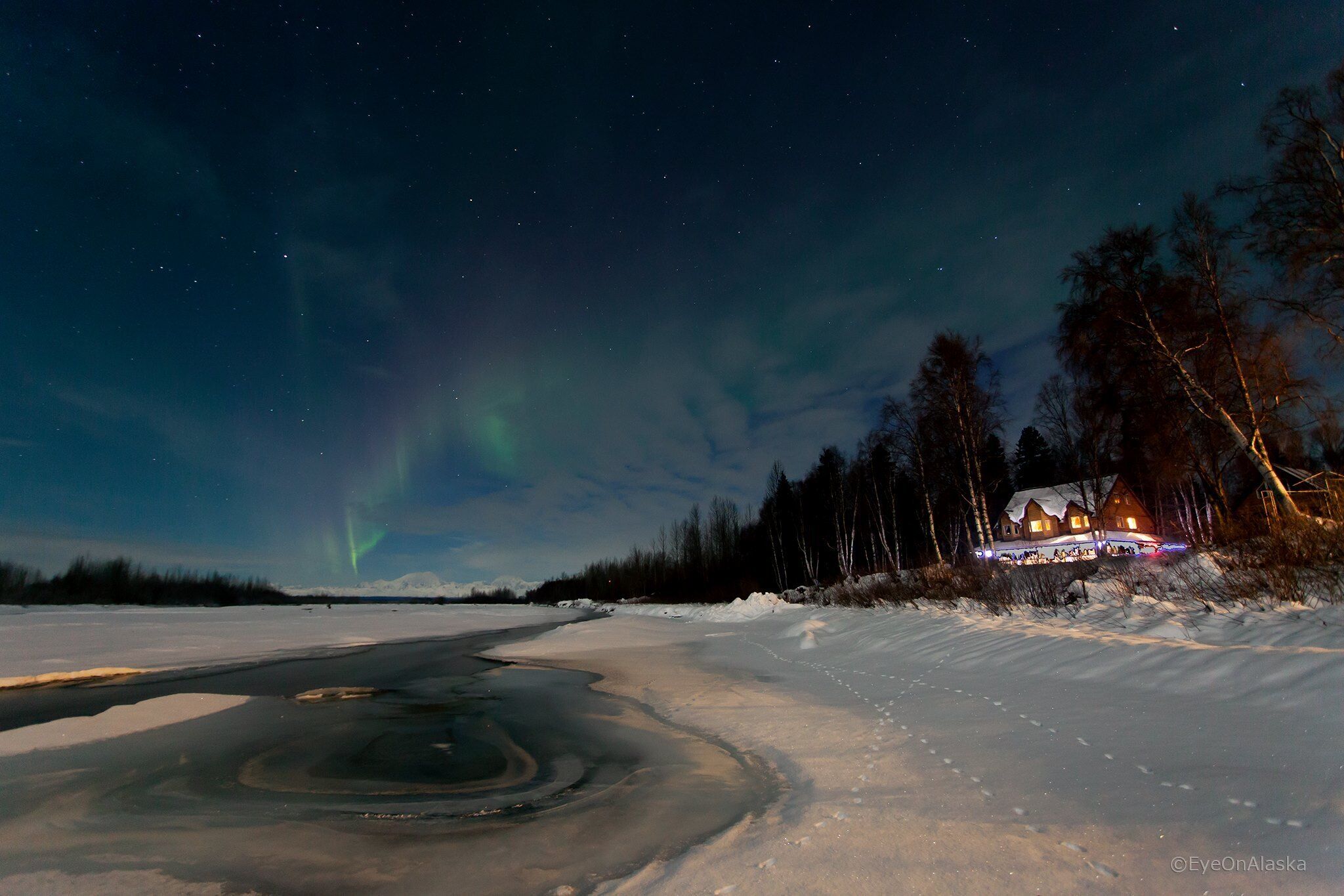 Un paisaje nevado con una casa iluminada por luces, rodeada de árboles y una masa de agua congelada.