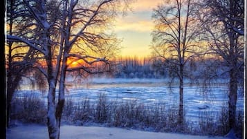 A winter landscape with a frozen river, leafless trees, and a sunset in the background.