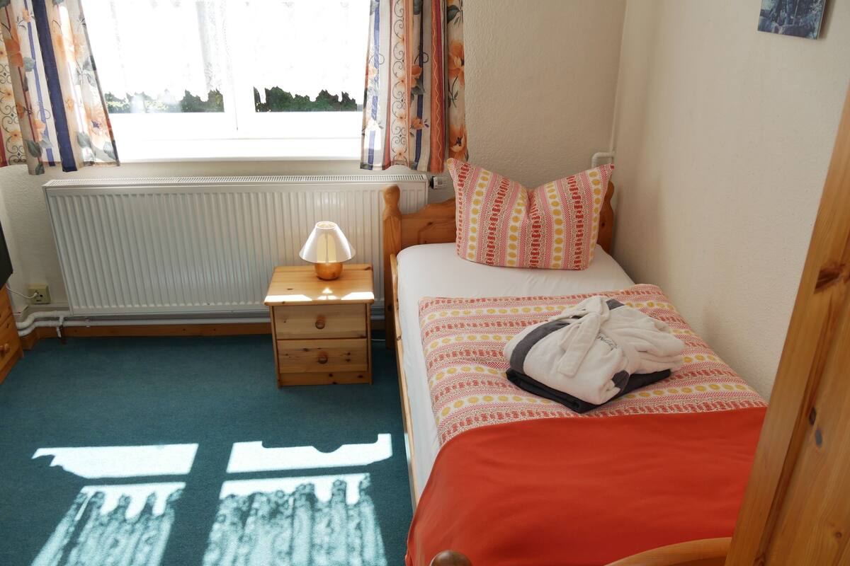 A bedroom with a bed, a bedside table with a lamp, a radiator, and a window with curtains.