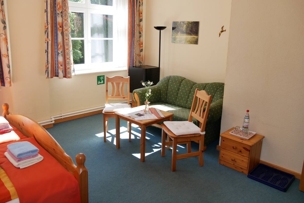 A cozy room with a bed, a green sofa, a wooden chair, a table with papers, a lamp, and a small cabinet.