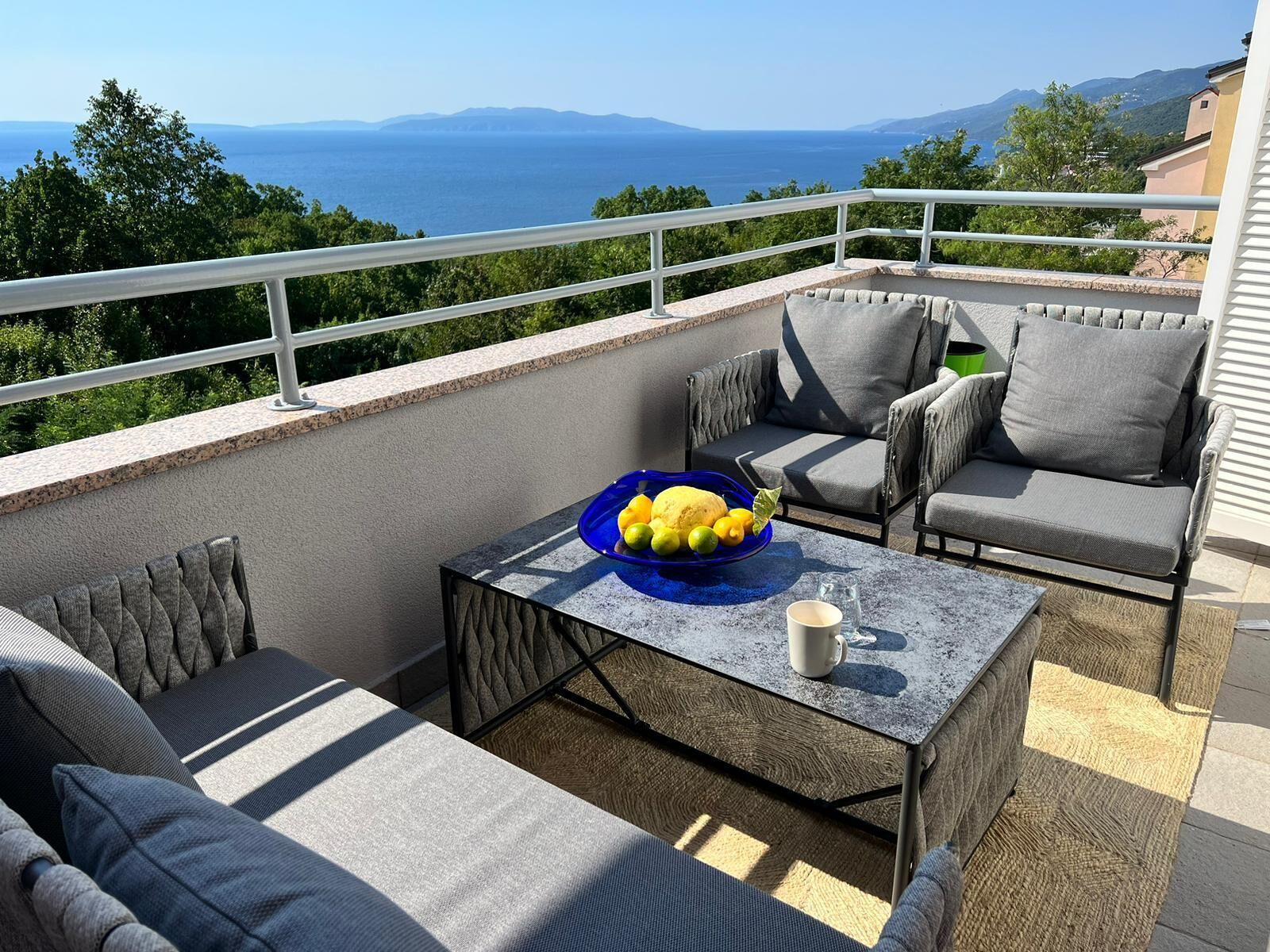 Ein Balkon mit Korbmöbeln, einem Tisch mit einer blauen Fruchtschale und einem Glas Wasser.