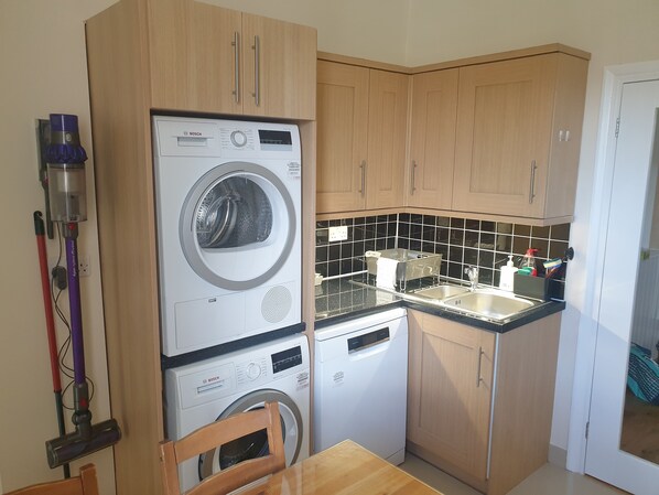 A compact kitchen with a Bosch washing machine, a dishwasher, wooden cabinets, a black tiled backsplash, and a sink with various kitchen items.