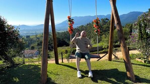 Un columpio de madera con flores, una persona sentada y una vista de montañas y una casa al fondo.