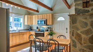 A kitchen with wooden cabinets, a dining table with chairs, a refrigerator, and a stone wall. - Firefly Cottage | Pet-Friendly, Fire Pit, 8 Mins to DT AVL + Biltmore Pass (Asheville)