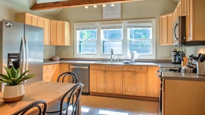 A kitchen with wooden cabinets, a stainless steel refrigerator, a dining table with chairs, and a window with blinds. - Firefly Cottage | Pet-Friendly, Fire Pit, 8 Mins to DT AVL + Biltmore Pass (Asheville)