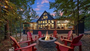 A cabin with a fire pit surrounded by red Adirondack chairs, set in a forested area.