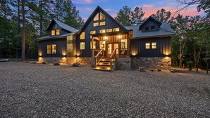 A large, dark wooden cabin with a stone foundation, a central staircase, and a gravel driveway.