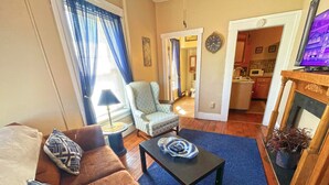 A living room with a brown sofa, patterned pillows, a black coffee table, and a blue rug. A light blue armchair and a small table with a lamp are also visible. - Nye's Place in Heart of Historic Roscoe Village (Coshocton)