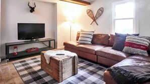A living room with a television on a stand, a sofa with cushions, a plaid rug, and a wooden chest of drawers. - Never Lonesome Hunter: Smart 4bd Getaway near Mtn (Hunter)