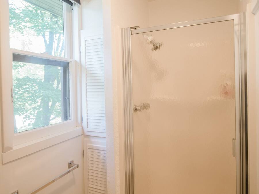 A bathroom with a glass shower enclosure, a window with blinds, and a white sink.
