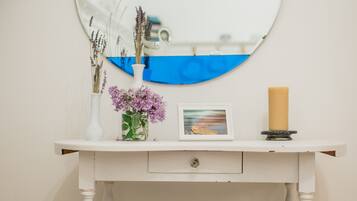 A white wooden vanity with a round mirror, a vase with purple flowers, a candle, and a framed picture.