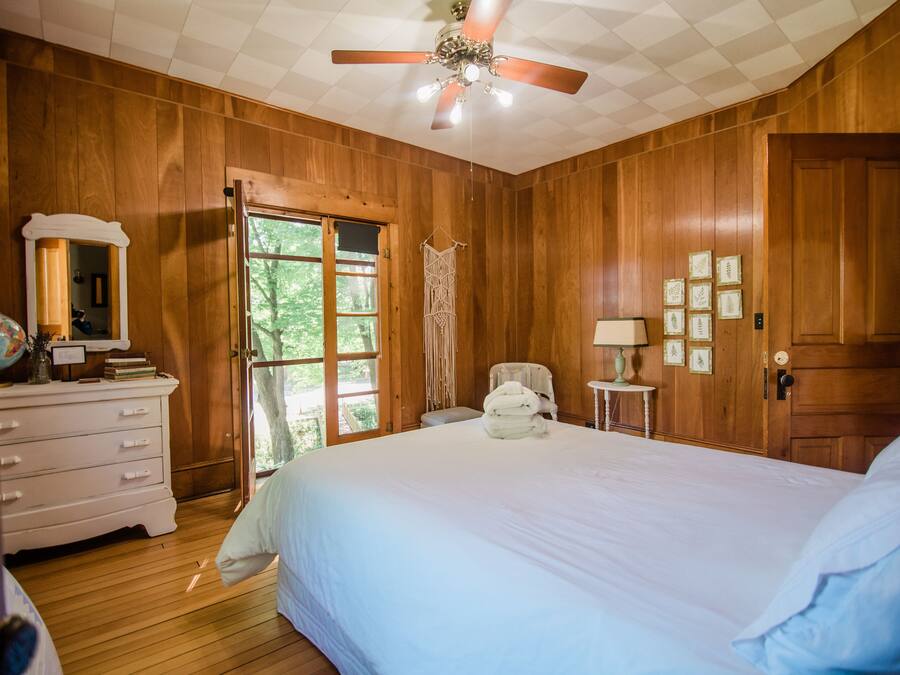 Une chambre avec des murs en bois, un grand lit, un ventilateur de plafond, une commode, une chaise et une porte.