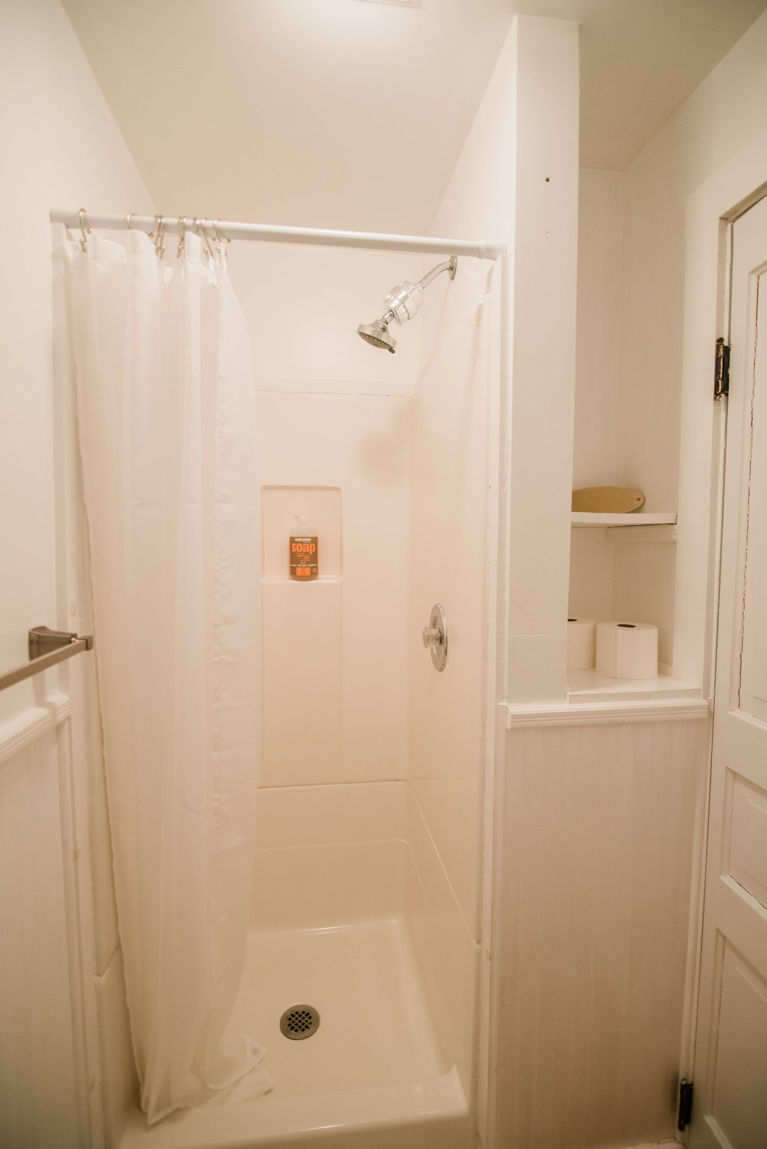 Une petite salle de bain blanche, équipée d’une douche, de toilettes et d’une étagère où sont rangés les articles de toilette.