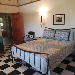 Ein Zimmer mit Schachbrettmuster-Boden, einem Bett mit gemusterten Betttextilien, einer Stehlampe, einem gerahmten Bild an der Wand und einer Tür zu einem anderen Raum.