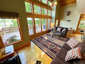 A cozy living room with a large window, wooden flooring, a sofa with patterned cushions, and a mounted deer head on the wall. - Hilltop Hideaway - Great views of Lake George (Bolton)