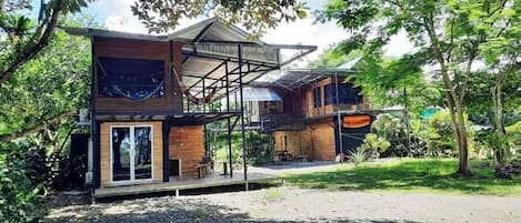A two-story wooden house with a balcony, surrounded by lush greenery.