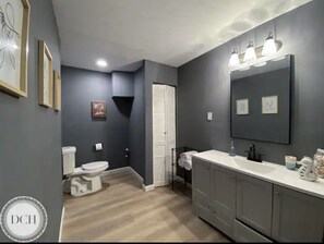 Bathroom features a white toilet, sink, and towel rack, with framed pictures on the wall and a door in the background.