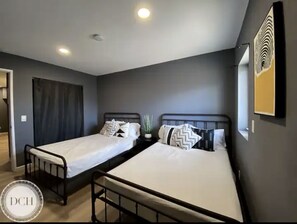 Two single beds with white mattresses and patterned pillows, set against a neutral gray backdrop.