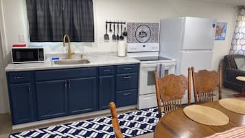 A compact kitchen with a microwave, sink, and stove. There are cabinets, a chair, and a dining table with placemats.