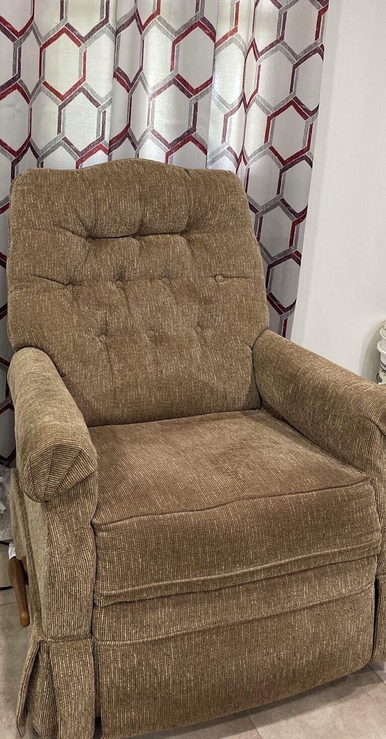 Un fauteuil marron, recouvert de tissu, avec un dossier et des accoudoirs capitonnés, placé contre un mur orné d’un rideau à motifs géométriques.