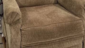 Un fauteuil marron, recouvert de tissu, avec un dossier et des accoudoirs capitonnés, placé contre un mur orné d’un rideau à motifs géométriques.