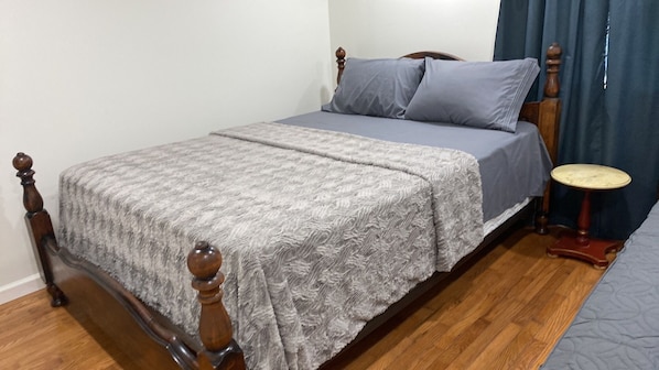 A single bed with a wooden frame and a grey bedspread, a small round table, and a blue curtain.