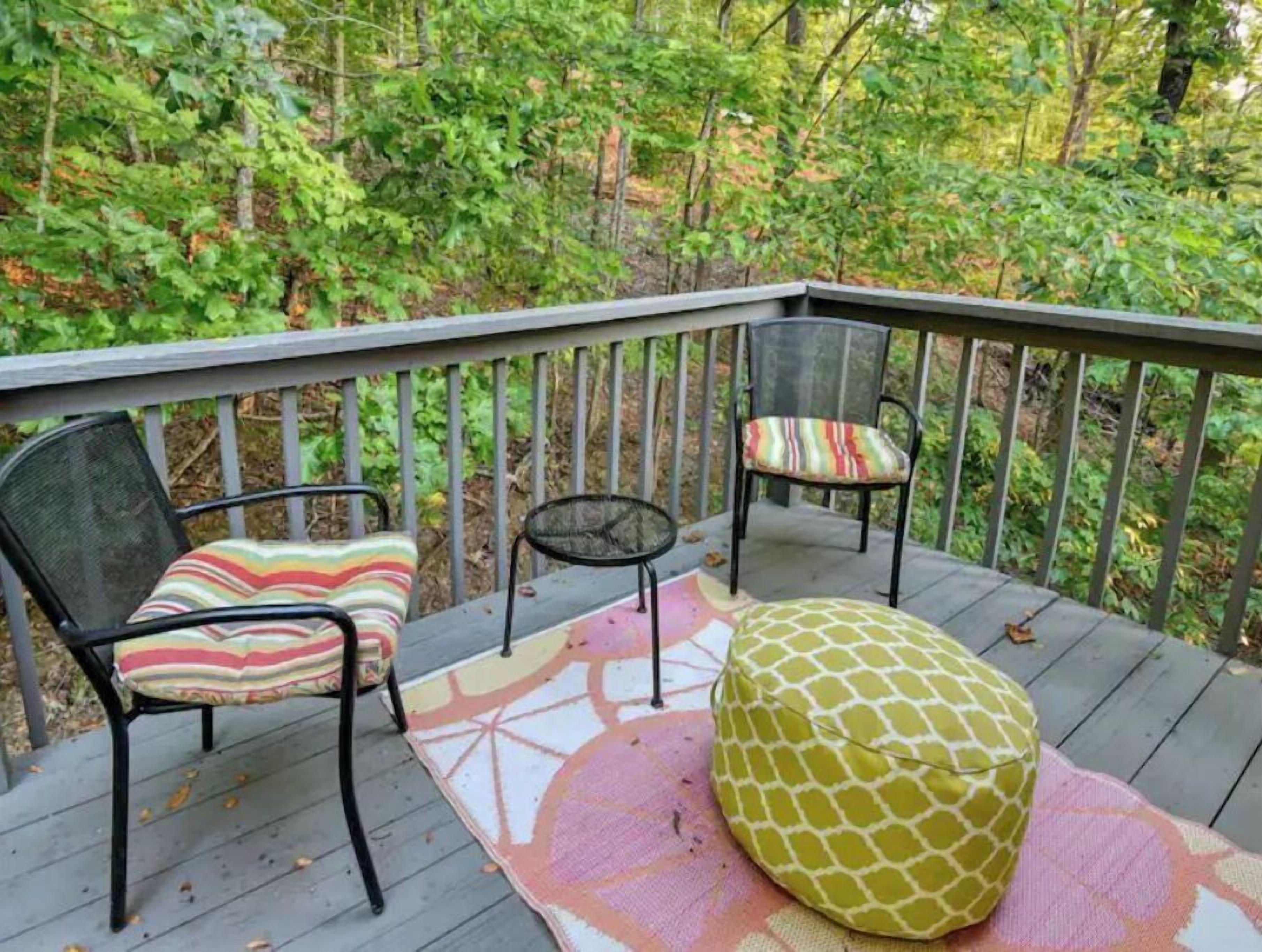 Una terraza de madera con dos sillas y un reposapiés redondo, rodeada de un bosque.