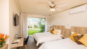 Habitación de hotel con dos camas, un escritorio con una computadora portátil, un ventilador de techo y vista a un jardín.