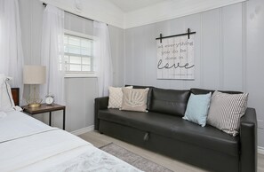 A bedroom with a bed, a nightstand with a lamp, a black leather sofa with pillows, a wall with a quote, and a window with curtains. - Cute, Cozy and Clean cottage by the Sea! (Freeport)