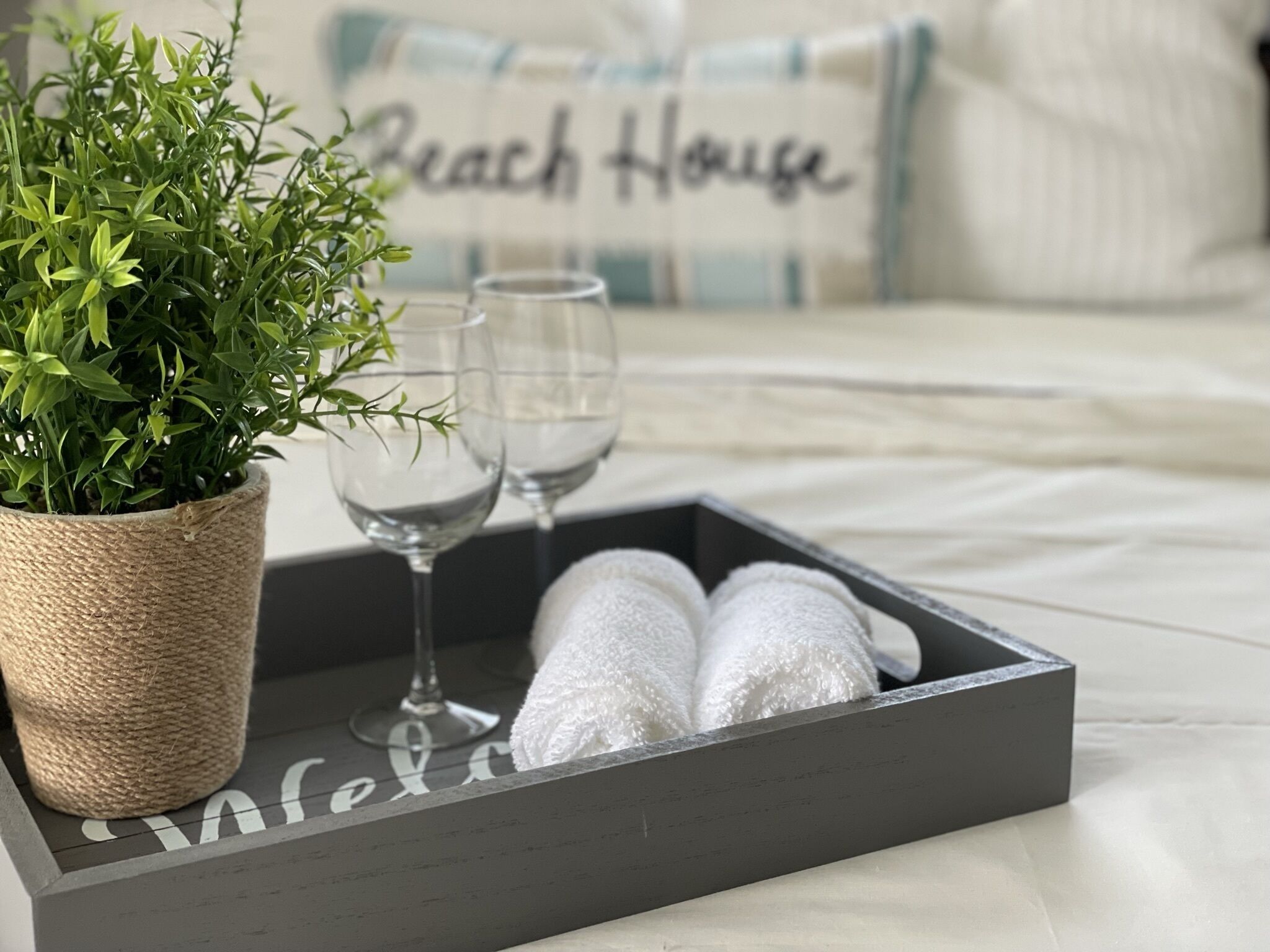 Una bandeja con dos toallas blancas y una planta en maceta sobre una cama con un cojín que dice 'Beach House'.