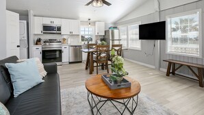 A modern kitchen with white cabinets, stainless steel appliances, and a dining area with wooden chairs. - Cute, Cozy and Clean cottage by the Sea! (Freeport)