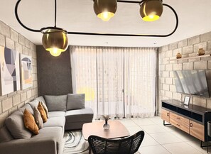 A modern living room with a sectional sofa, a coffee table, a dining table, and a TV mounted on the wall.