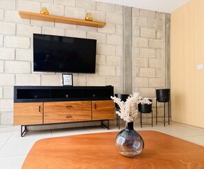 Un salon moderne avec un téléviseur fixé sur un mur en briques, une console en bois, deux chaises noires et un vase contenant des fleurs séchées.