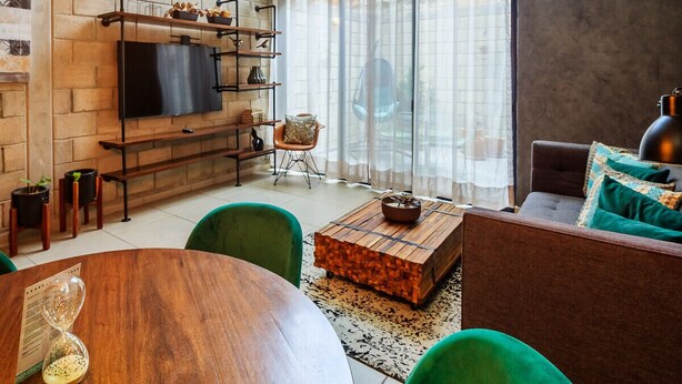 Un salon moderne avec un mur en briques, un téléviseur à écran plat, une table basse en bois, des fauteuils verts et un canapé agrémenté de coussins à motifs.