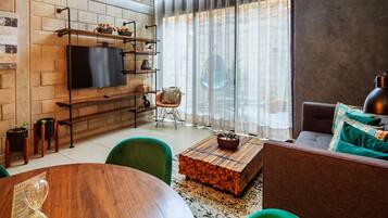 Una sala de estar moderna con pared de ladrillo, un televisor de pantalla plana, una mesa de centro de madera, sillas verdes y un sofá con cojines estampados.